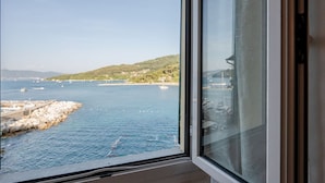 Vista costera a través de una ventana, con una costa rocosa, barcos y una colina distante.