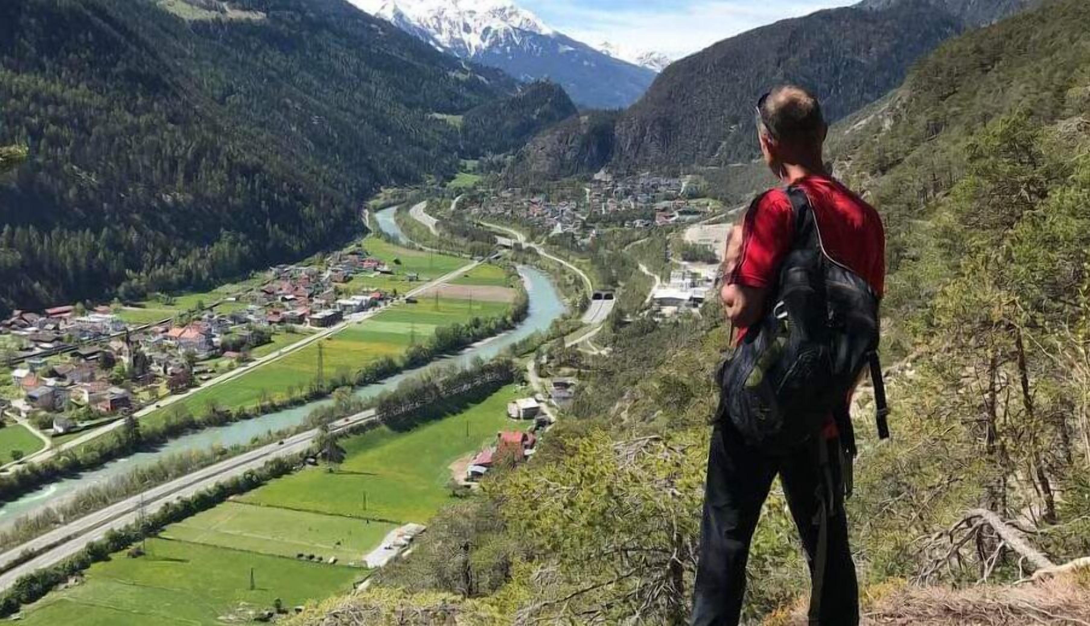 Eine Person blickt über ein Tal mit einem Fluss, Häusern und Bergen.