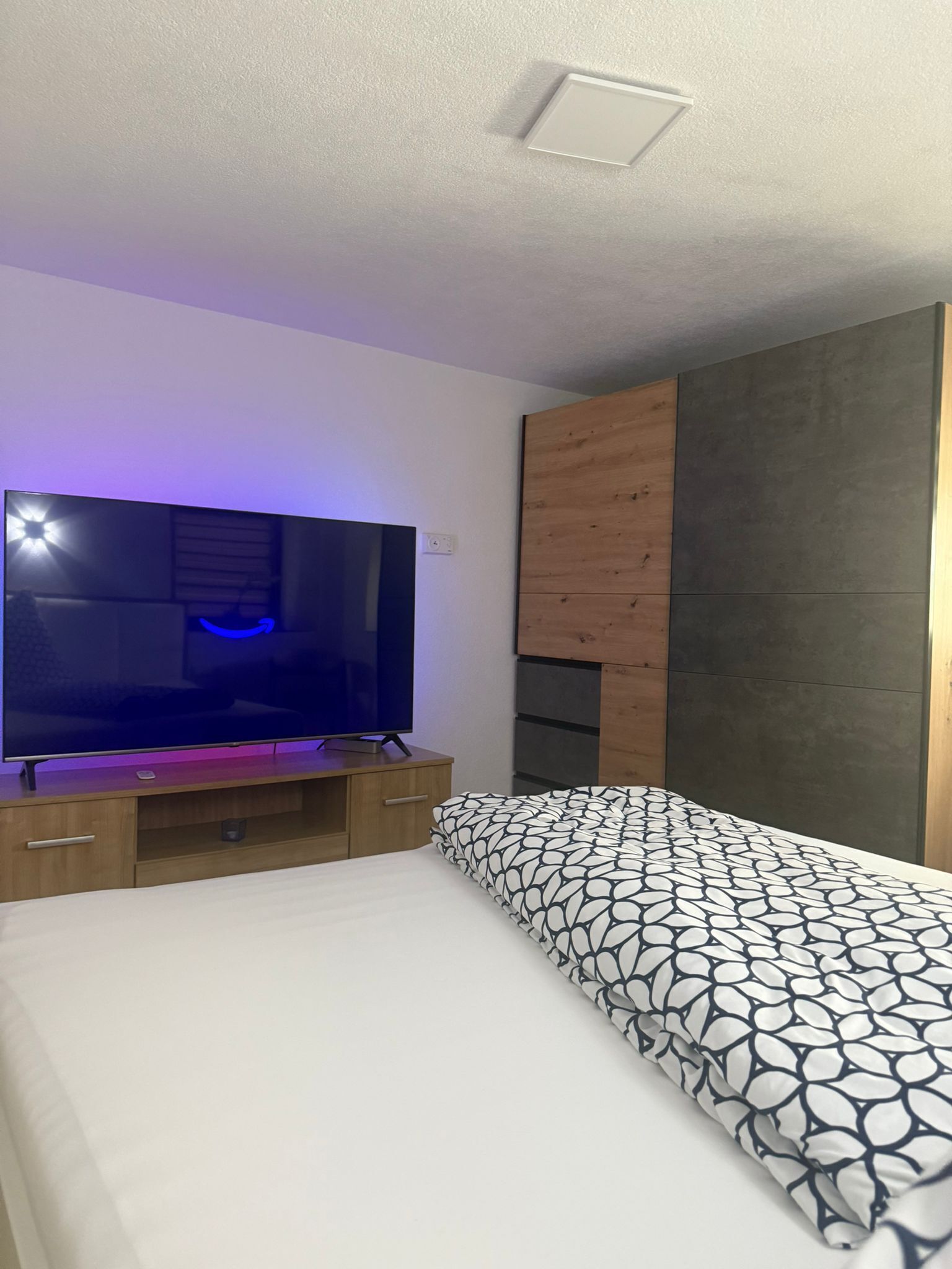 Une chambre à coucher avec un lit, un téléviseur posé sur un meuble en bois et une armoire dont le design combine du bois et des panneaux foncés.