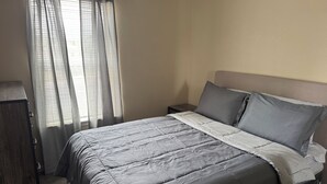 Una cama bien hecha con colcha gris, una mesita de noche de madera y una ventana con cortinas traslúcidas.