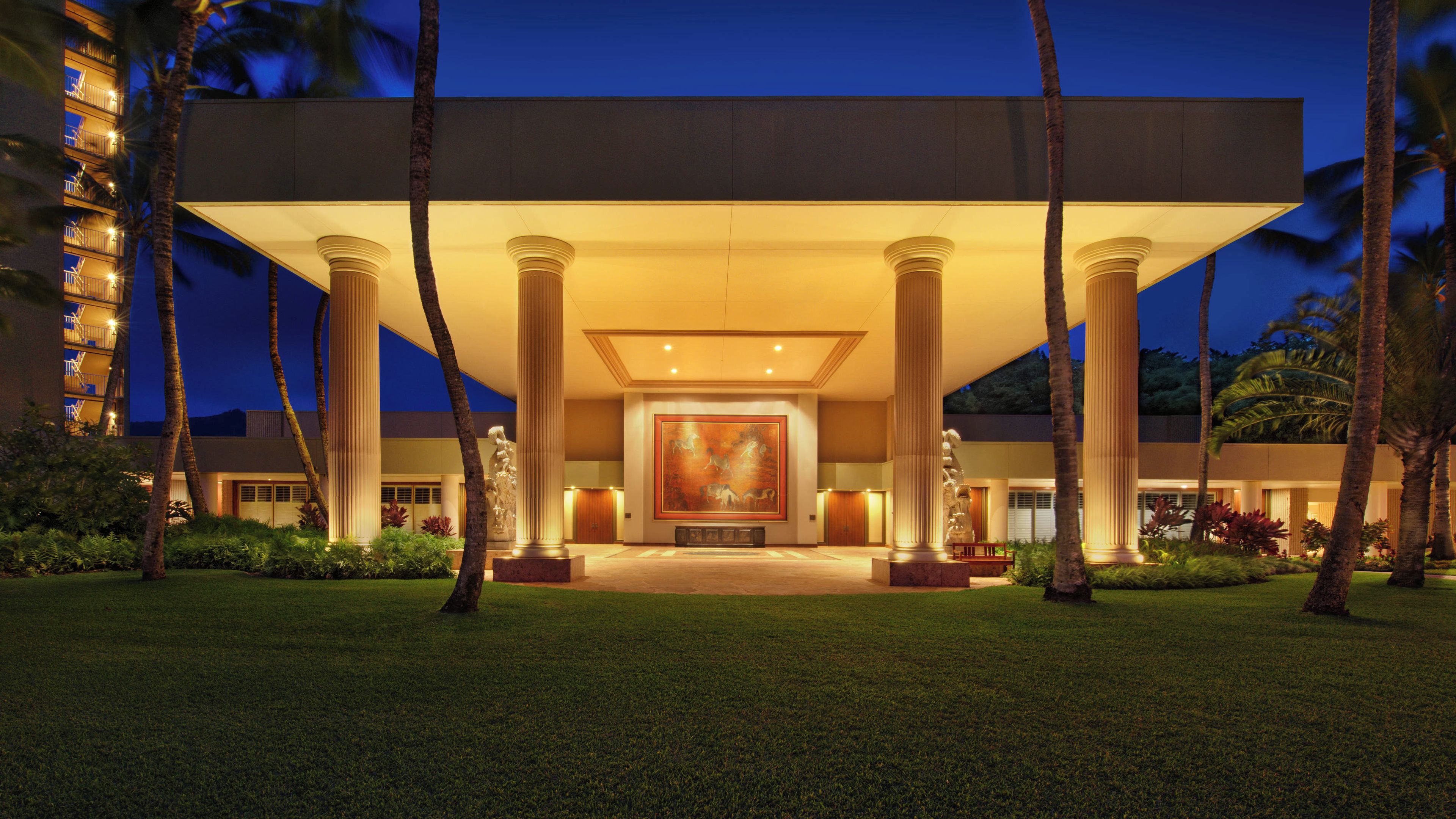 A grand hotel entrance with a large central mural, symmetrical columns, and a well-maintained lawn.