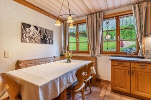 A dining area with a table set for four, wooden chairs, a painting on the wall, and a view of a garden through large windows. - Apartment 'Igelbau' with Mountain View, Shared Pool and Wi-Fi (Wolfach)