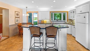 Une cuisine moderne avec des meubles blancs, un îlot central doté de tabourets et une grande fenêtre offrant une vue sur la verdure.