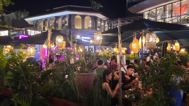 Un área de comedor al aire libre con mesas y sillas, rodeada de plantas en macetas y luces colgantes.