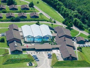An aerial view of a building complex with a central swimming pool, surrounded by multiple smaller buildings, a parking area, and green lawns.