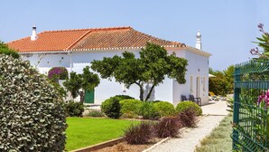 A white building with a red-tiled roof, surrounded by a well-maintained garden with various plants and a pathway leading to a seating area with lounge chairs. - Villa Caicco (Lagos)