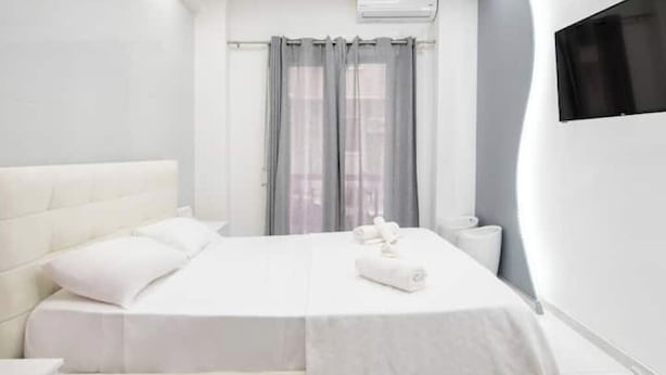 A modern hotel room with a large bed, a bedside table, a window with curtains, and a flat-screen TV mounted on the wall. - Estia apartment (Athina)