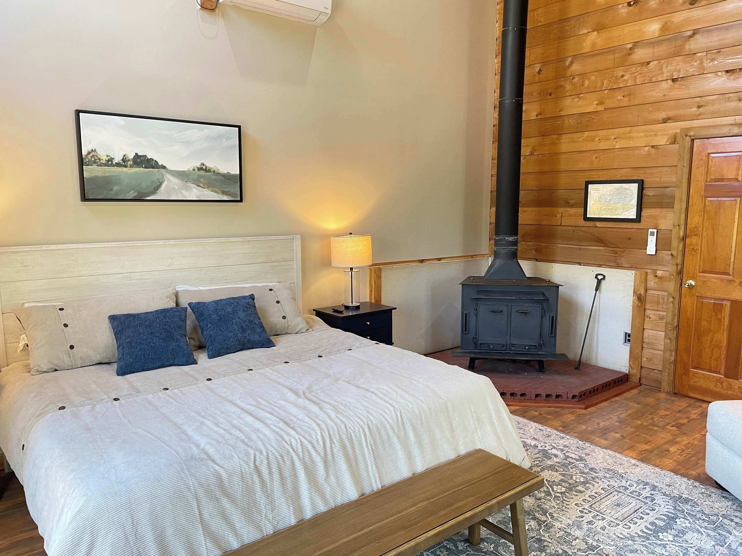 Une chambre à coucher comprenant un lit, une table de chevet, une lampe, un banc en bois, un climatiseur fixé au mur, un tableau encadré, une porte en bois et un poêle à bois.