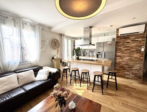 Un salon moderne avec un canapé en cuir noir, une table basse en bois et un coin cuisine équipé de tabourets de bar.