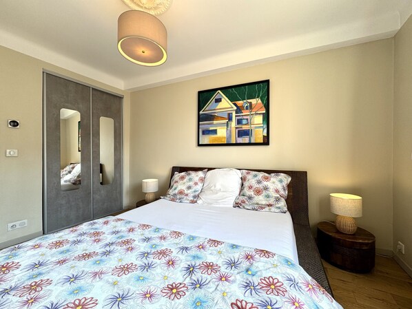 A bedroom with a bed, a nightstand, a lamp, a mirror, a painting, and a door. - Balcon de la Providence (Niza)