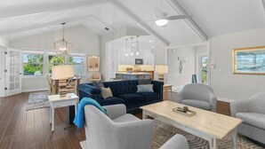 A spacious living area with a dark blue sofa, grey armchairs, and a wooden coffee table. A kitchen is visible in the background, and there are large windows offering a view of greenery.