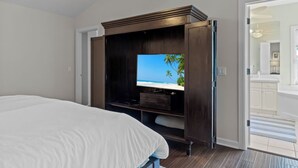 A bedroom with a bed, a TV on a wooden stand, and a view of the beach through the window.