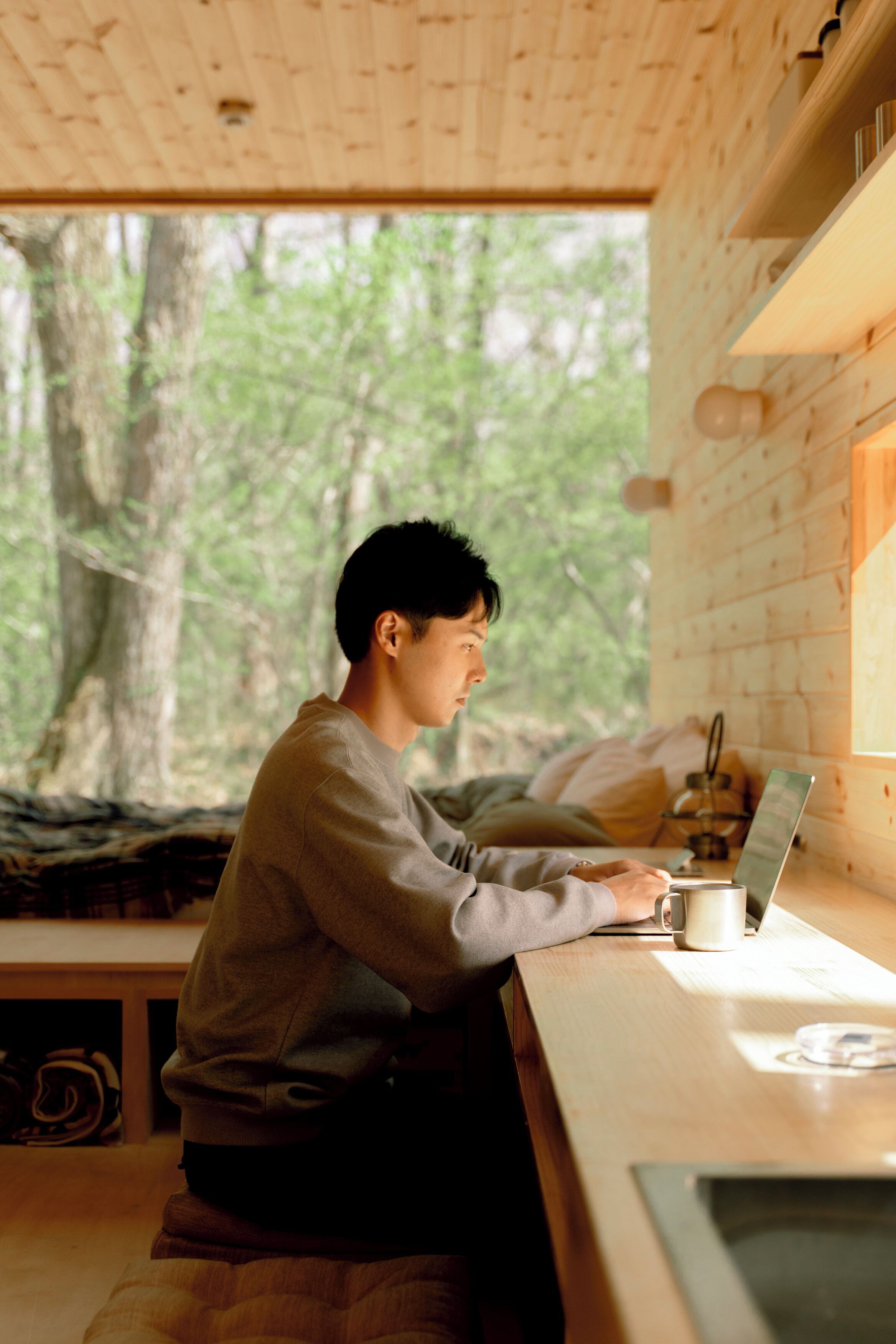 Une personne est assise à une table en bois, devant un ordinateur portable, dans une pièce d’où l’on peut voir des arbres à travers une fenêtre.
