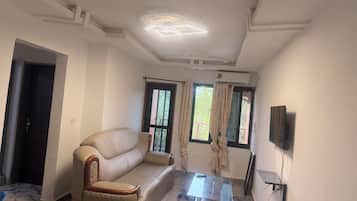 Una sala de estar con un sofá, una mesa de centro de vidrio, un televisor de pantalla plana montado en la pared y un ventanal con cortinas.