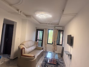 Una sala de estar con un sofá, una mesa de centro de vidrio, un televisor de pantalla plana montado en la pared y un ventanal con cortinas.