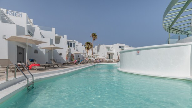 A swimming pool with clear water, surrounded by white buildings with balconies and umbrellas. - 'Kikere Puerto del Carmen' Holidays with Shared Pool, Wi-Fi and Air Conditioning (Puerto del Carmen)