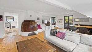 A modern living room with a wooden coffee table, a sofa with cushions, a fireplace, a dining area with chairs, and a kitchen in the background. - Spacious Allgäu loft | maisonette style (Nesselwang)
