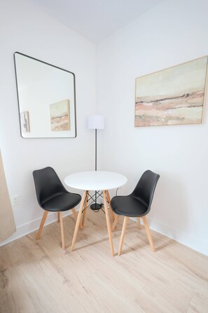 Una habitación con una mesa redonda blanca, dos sillas negras, una lámpara de pie y dos obras de arte enmarcadas en las paredes.