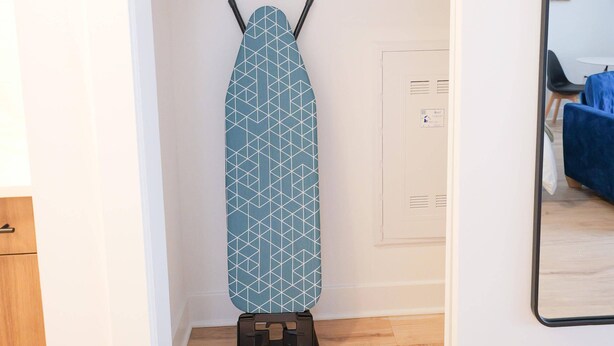 Una habitación con piso de madera, una puerta blanca, una tabla de planchar azul con diseño geométrico, un estante pequeño con un planchador azul, y un espejo en la pared.