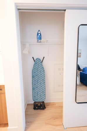 A room with a wooden floor, a white door, a blue ironing board with a geometric pattern, a small shelf with a blue iron, and a mirror on the wall.
