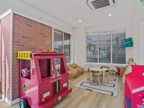 Una habitación con área de juegos con camión de bomberos rojo, ventana con vista a un edificio y televisor montado en la pared.