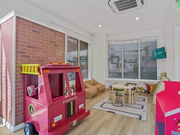 Una habitación con área de juegos con camión de bomberos rojo, ventana con vista a un edificio y televisor montado en la pared.