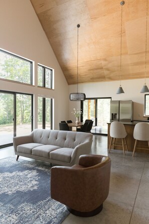 En moderne stue med hvælving, store vinduer, sofa, sofabord og et køkkenområde med barstole.