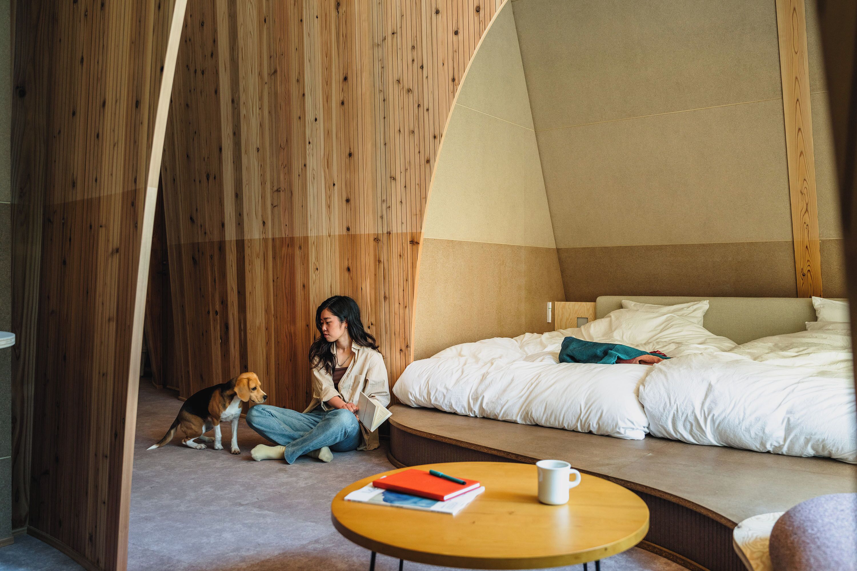 Una habitación con una cama, piso de madera, una mesa redonda con un libro y una taza, y un perro.