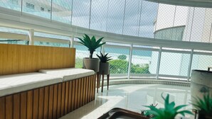 A modern reception area with a wooden bench, potted plants, and large windows offering a view of the cityscape. - CM 304 - luxo conforto em frente ao mar  (Cabo Frio)