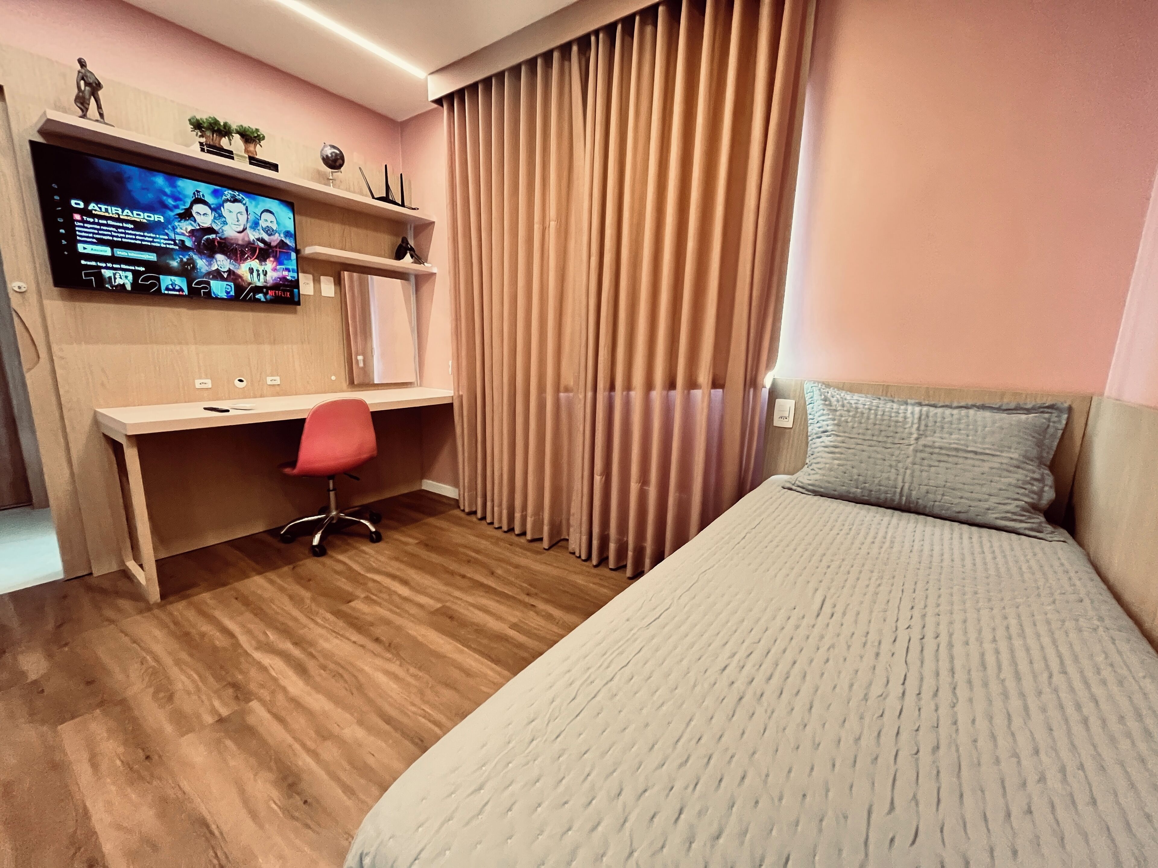 Quarto de hotel com cama, escrivaninha com cadeira, televisão na parede e janela com cortinas.