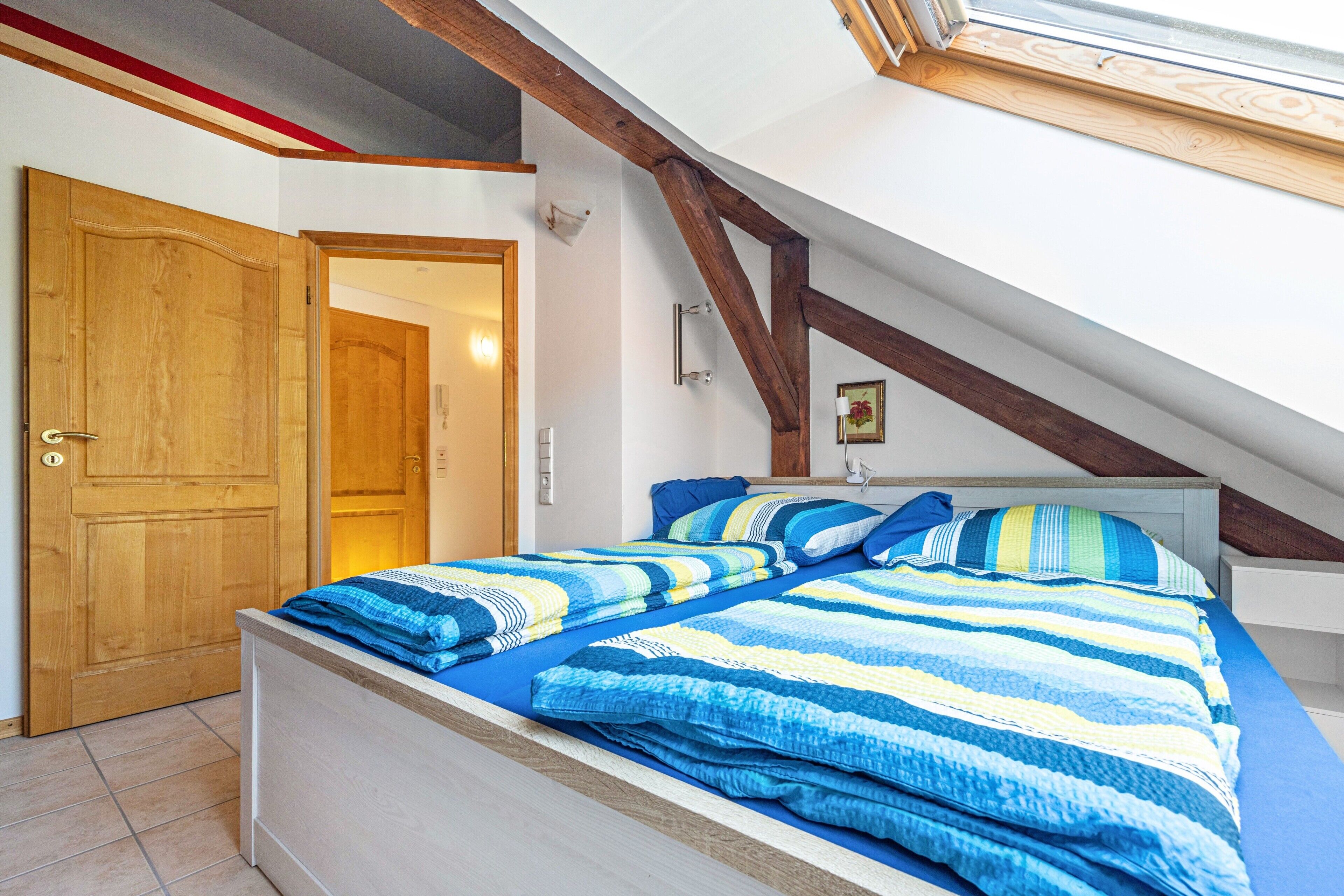 A bedroom with a sloped ceiling, a wooden door, a bed with striped bedding, and a white wall with a picture.