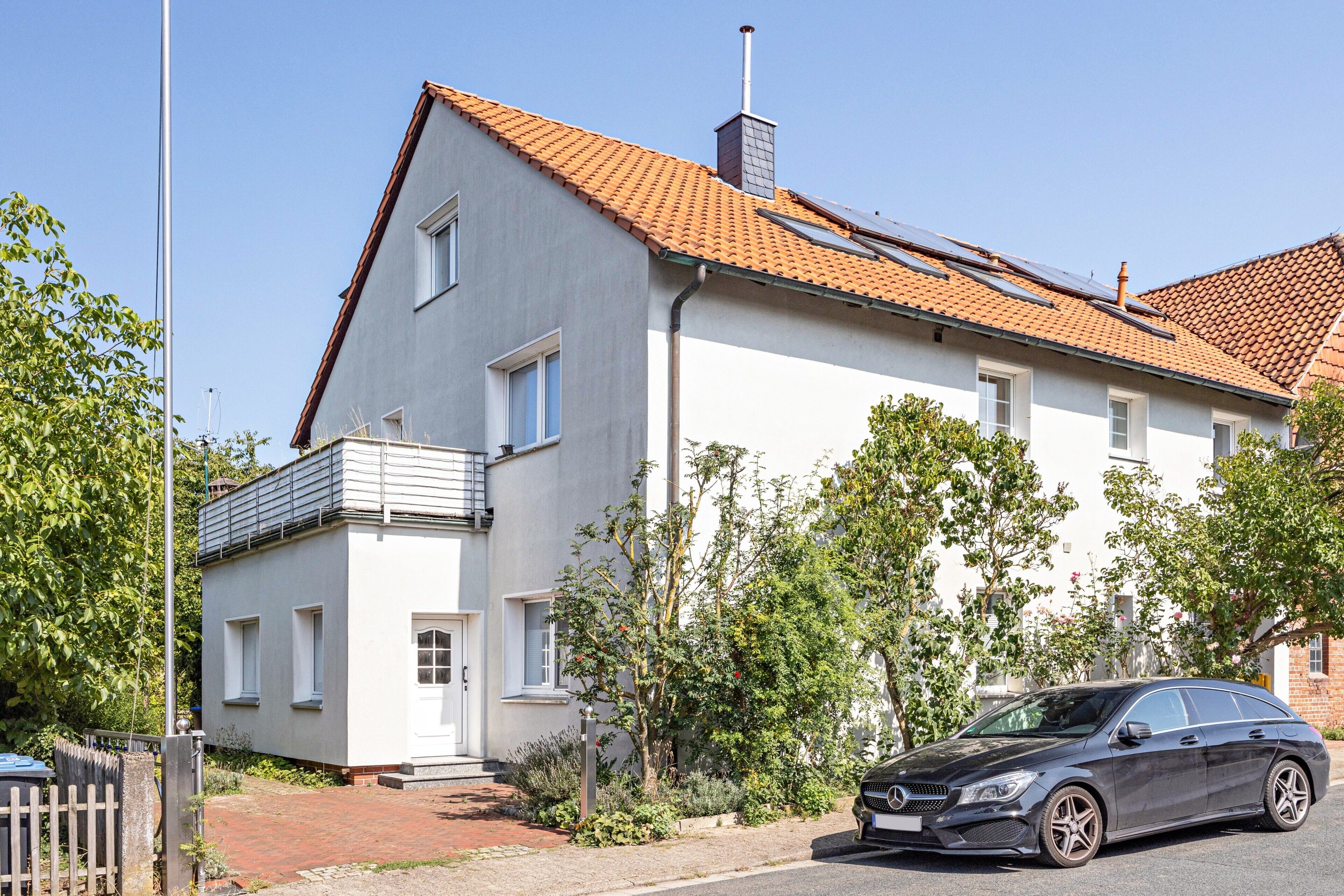 Une maison à deux étages avec un toit recouvert de tuiles rouges, une voiture garée dans l’allée et un jardin où poussent différentes espèces de plantes.