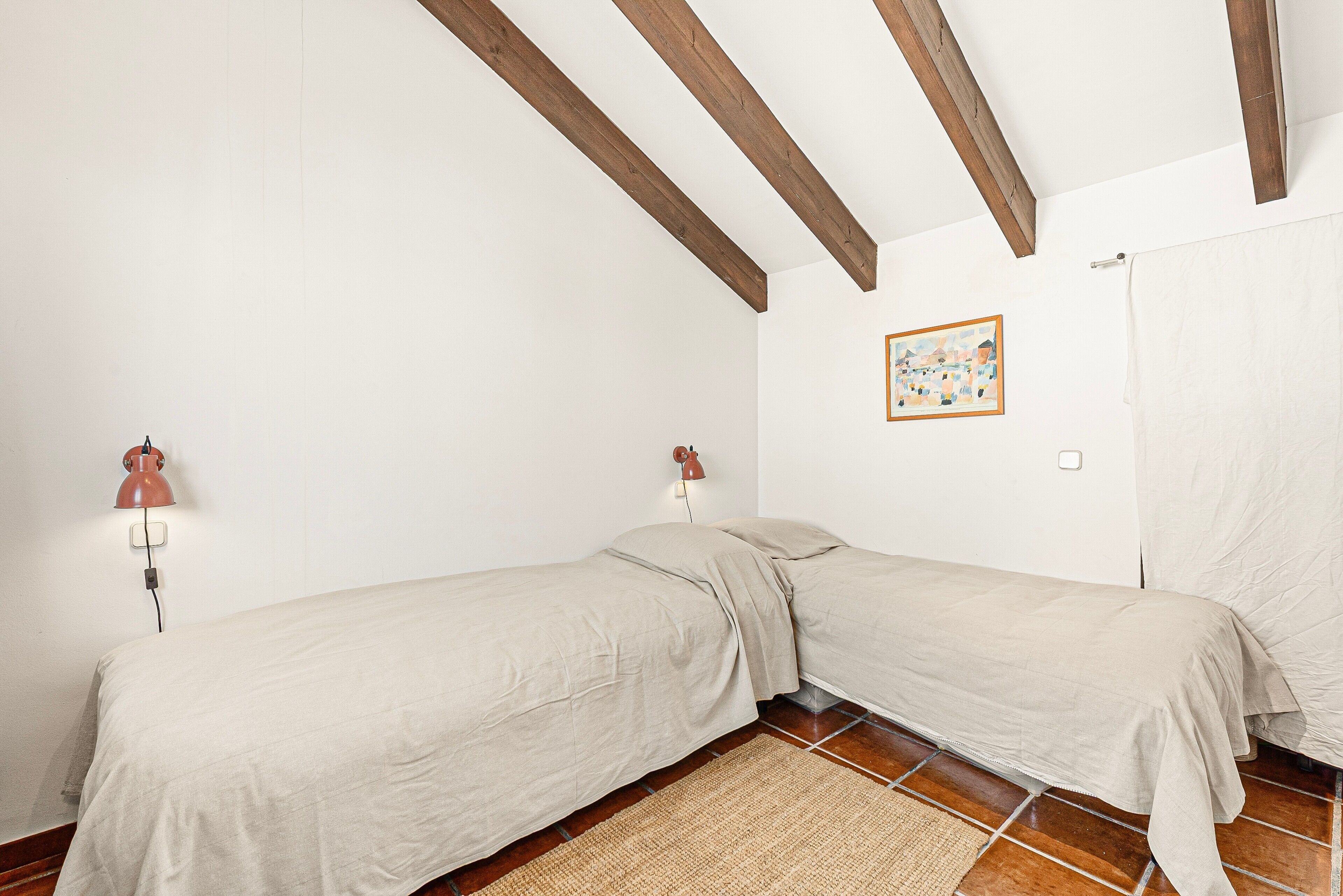 A room with two single beds, wooden beams on the ceiling, a wall-mounted lamp, and a tiled floor.
