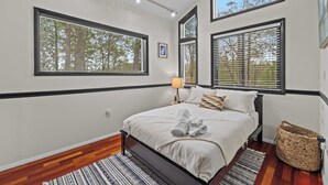 A bedroom with a large bed, a ceiling fan, and a view of trees through the windows. - Modern Lake View, Firepit, Hot Tub, Lake Views! (Tobyhanna)