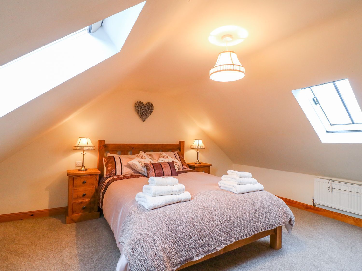 A cozy attic bedroom with a sloped ceiling, a bed with a headboard, two bedside lamps, a heart-shaped wall decoration, and a skylight.