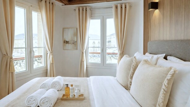A hotel room with a bed, towels, toiletries, and a view of buildings through the windows. - 841 Polytech (Paris)