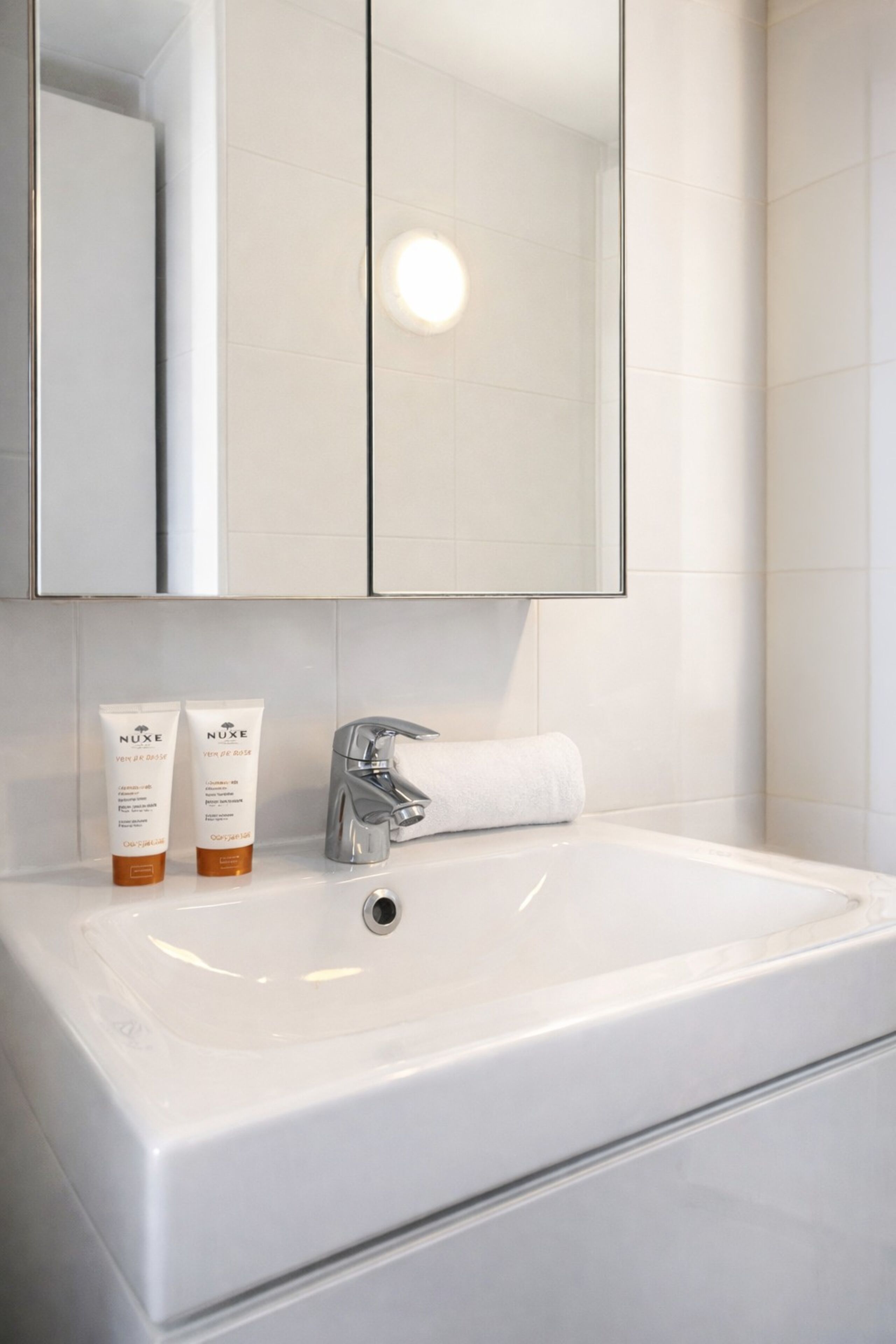 A bathroom with a white sink, two Nuxe skincare tubes, a towel, and a mirror above.