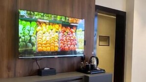A modern hotel room with a large flat-screen TV mounted on a wooden wall, a small black cabinet below, and a waste bin.