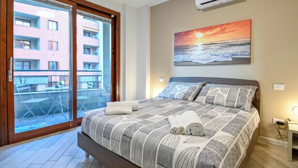 A bedroom with a large bed, a painting of a sunset, and a view of a building through a glass door. - [10min BGY Airport] Bilo Chic: garage + A/C (Seriate)