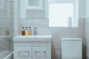 Un baño con un lavamanos blanco, tres botellas en la encimera, un inodoro y una ventana.