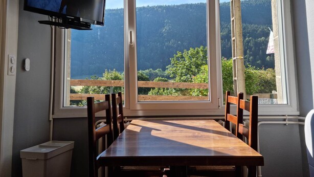 Ein Essbereich mit Tisch und Stühlen, einem an der Wand befestigten Fernseher und großen Fenstern mit Blick auf eine bewaldete Berglandschaft.