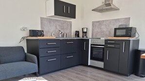 Eine kompakte Küche mit schwarzen Schränken, einer Mikrowelle, einem Herd und einer Kaffeemaschine. Ein graues Sofa und ein kleiner Tisch mit einer Fernbedienung sind ebenfalls vorhanden.