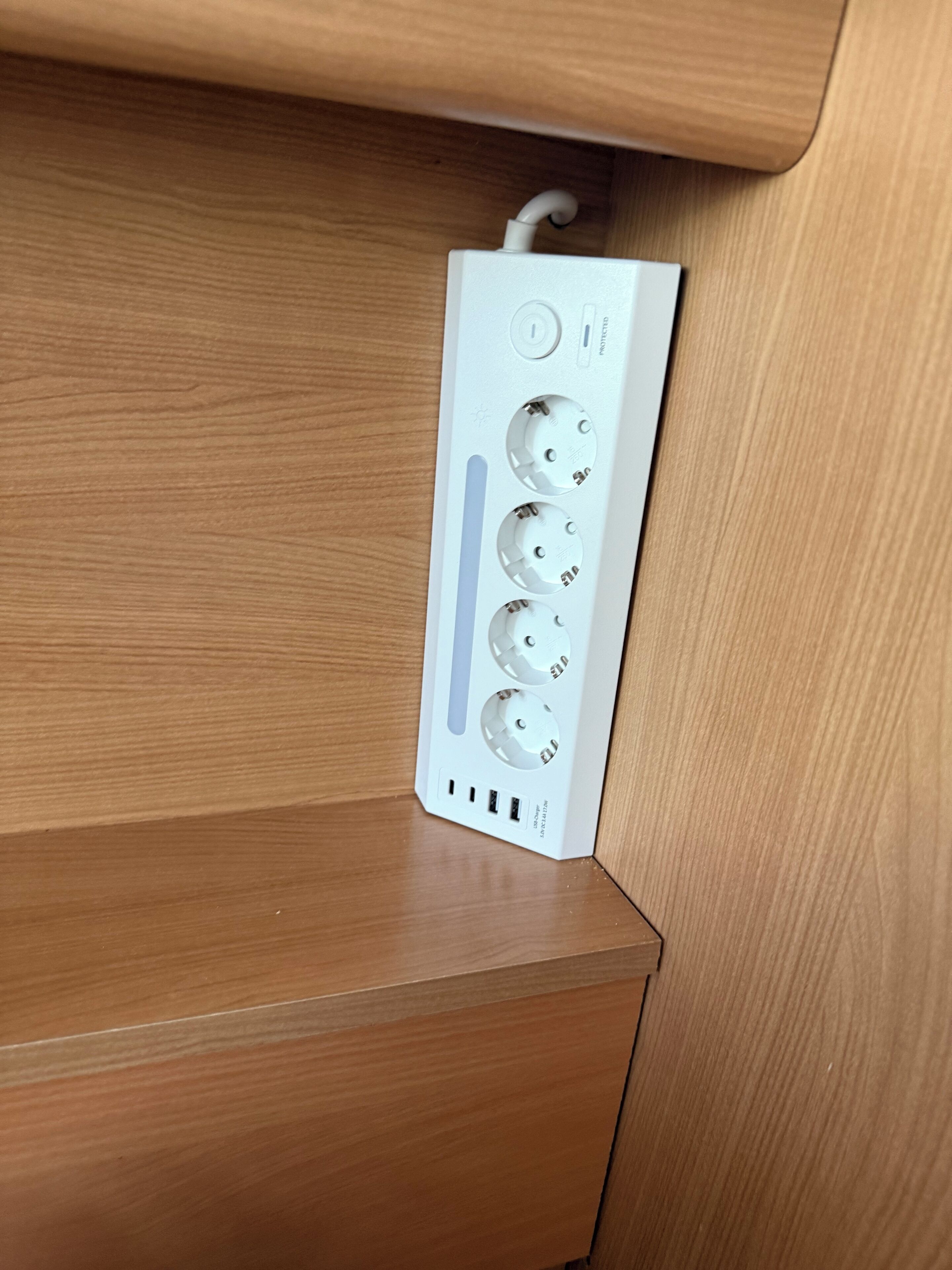 Tome de eletricidade branco com várias tomadas e portas USB, fixado numa superfície de madeira.