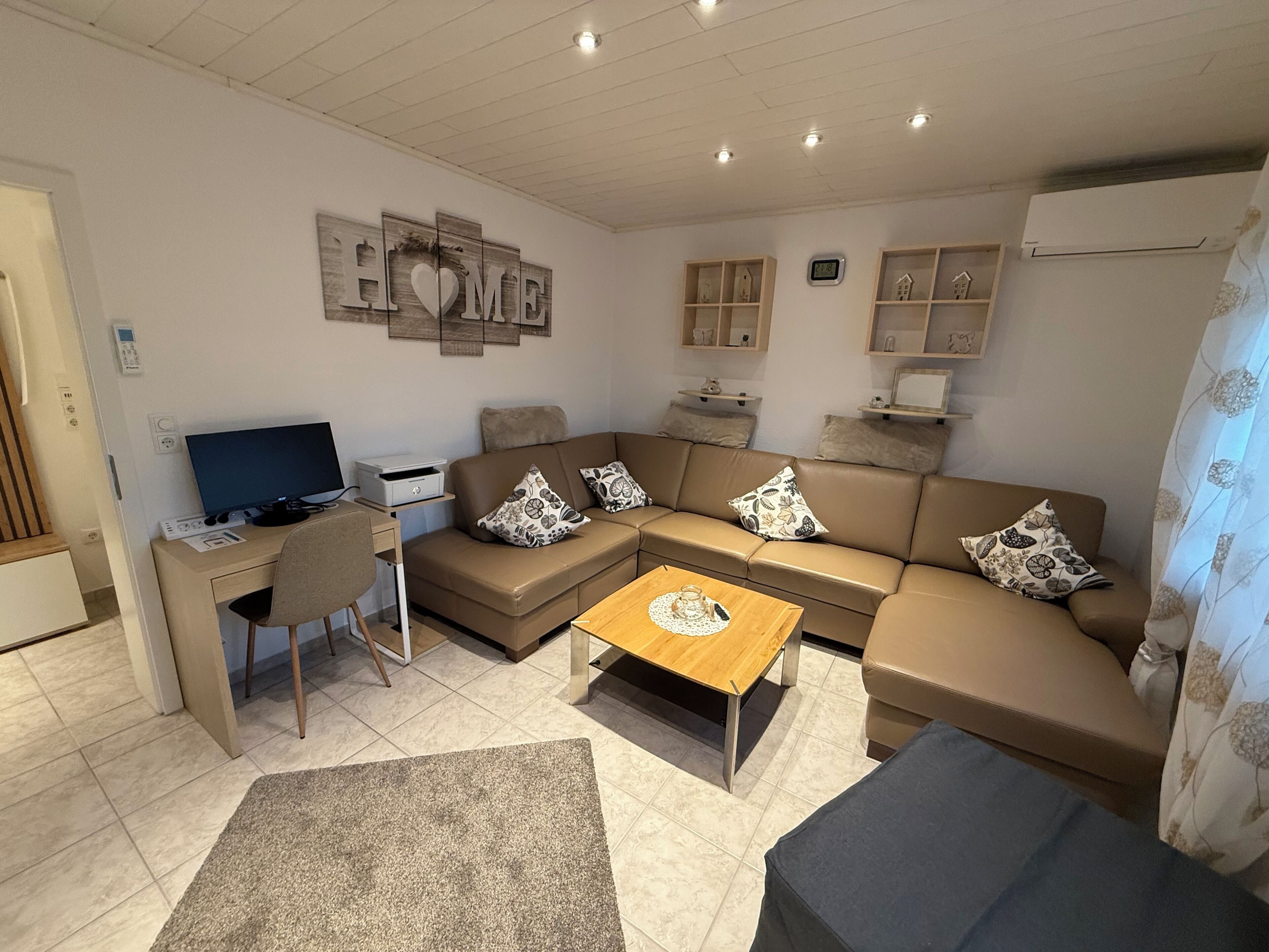 A modern living room with a sofa, coffee table, and a desk with a computer. There are decorative pillows and a wall art piece that reads "HOME".