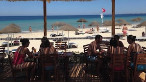 Un restaurante frente a la playa con terraza al aire libre y techo de paja, con vistas a la playa.