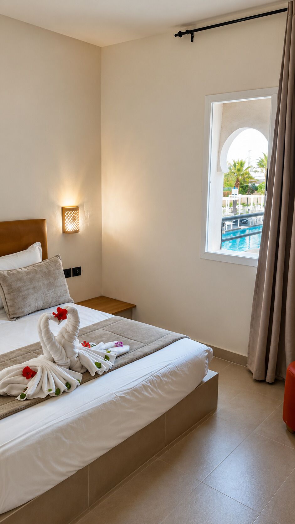 Une chambre d’hôtel avec un lit, une table de chevet, une lampe fixée au mur, une fenêtre donnant sur une piscine et un rideau.