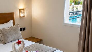 Une chambre d’hôtel avec un lit, une table de chevet, une lampe fixée au mur, une fenêtre donnant sur une piscine et un rideau.