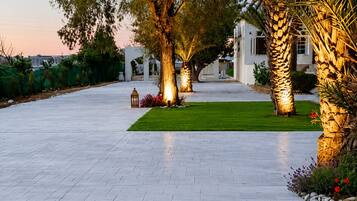 Un chemin pavé mène à un bâtiment blanc doté d’une entrée couverte, le tout étant entouré d’arbres et de plantes tropicales.
