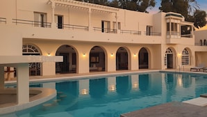 Una zona junto a la piscina con una pileta cristalina, un espacio de descanso techado y un edificio con múltiples arcos y balcones.