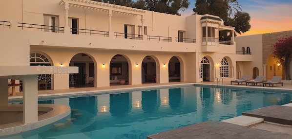 A poolside area with a clear pool, a covered seating area, and a building with multiple arches and balconies. - Hôtel Archipel (Monastir)